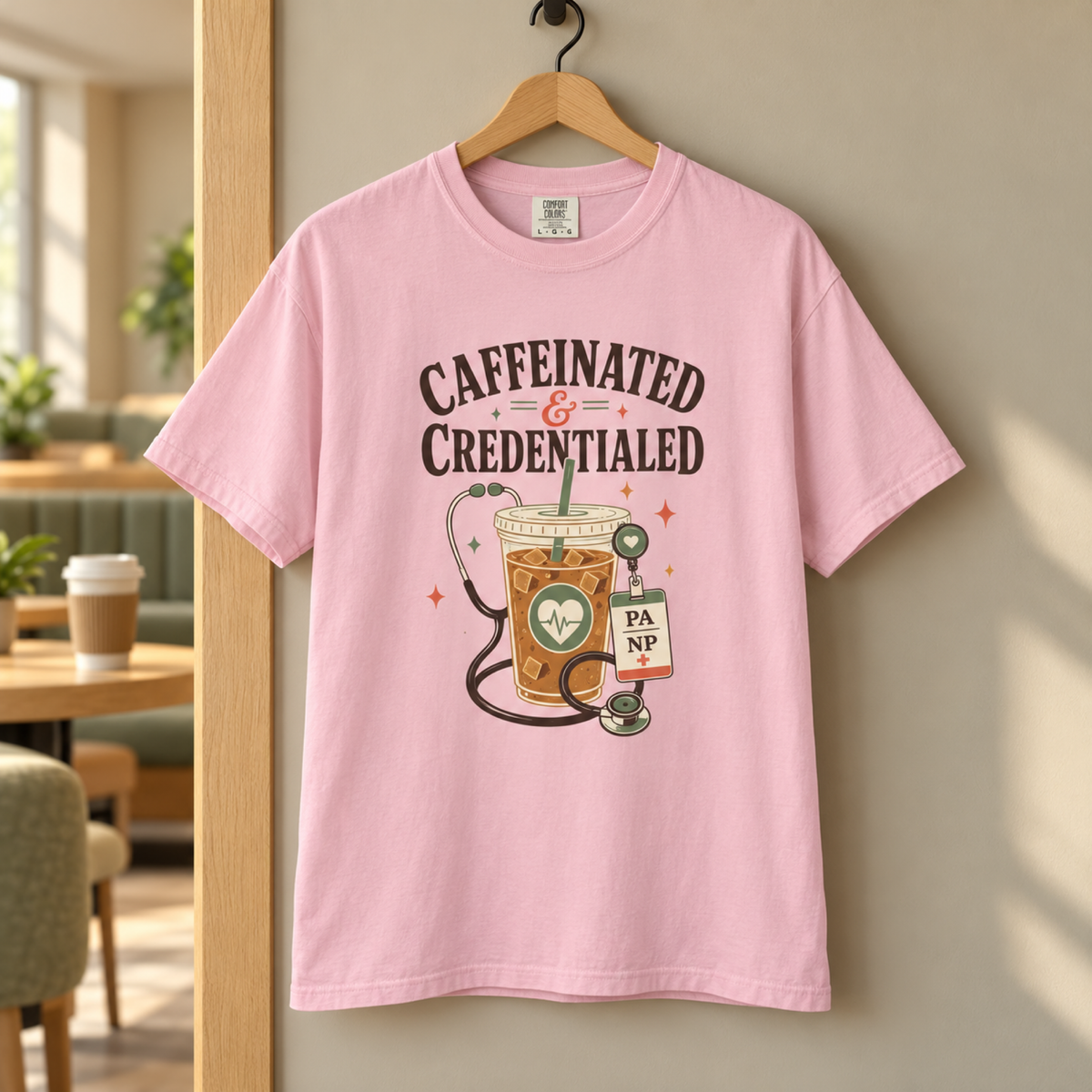 Blossom pink Caffeinated and Credentialed Comfort Colors shirt on a hanger in a warm staff coffee lounge