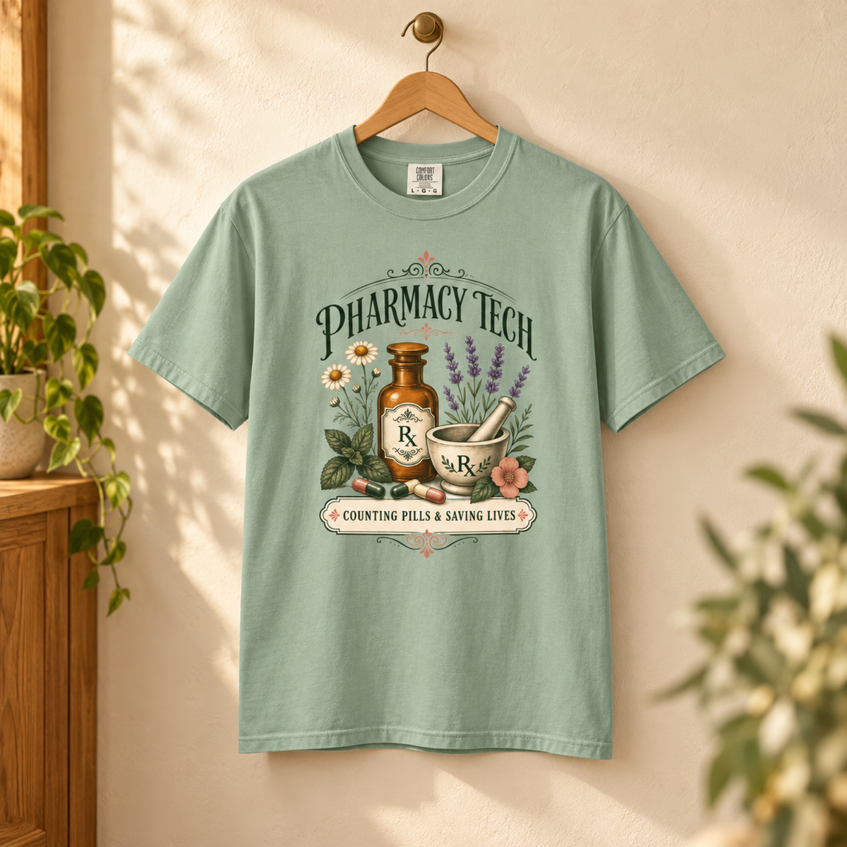 Bay green botanical Pharmacy Tech tee on a wooden hanger against a warm cream wall