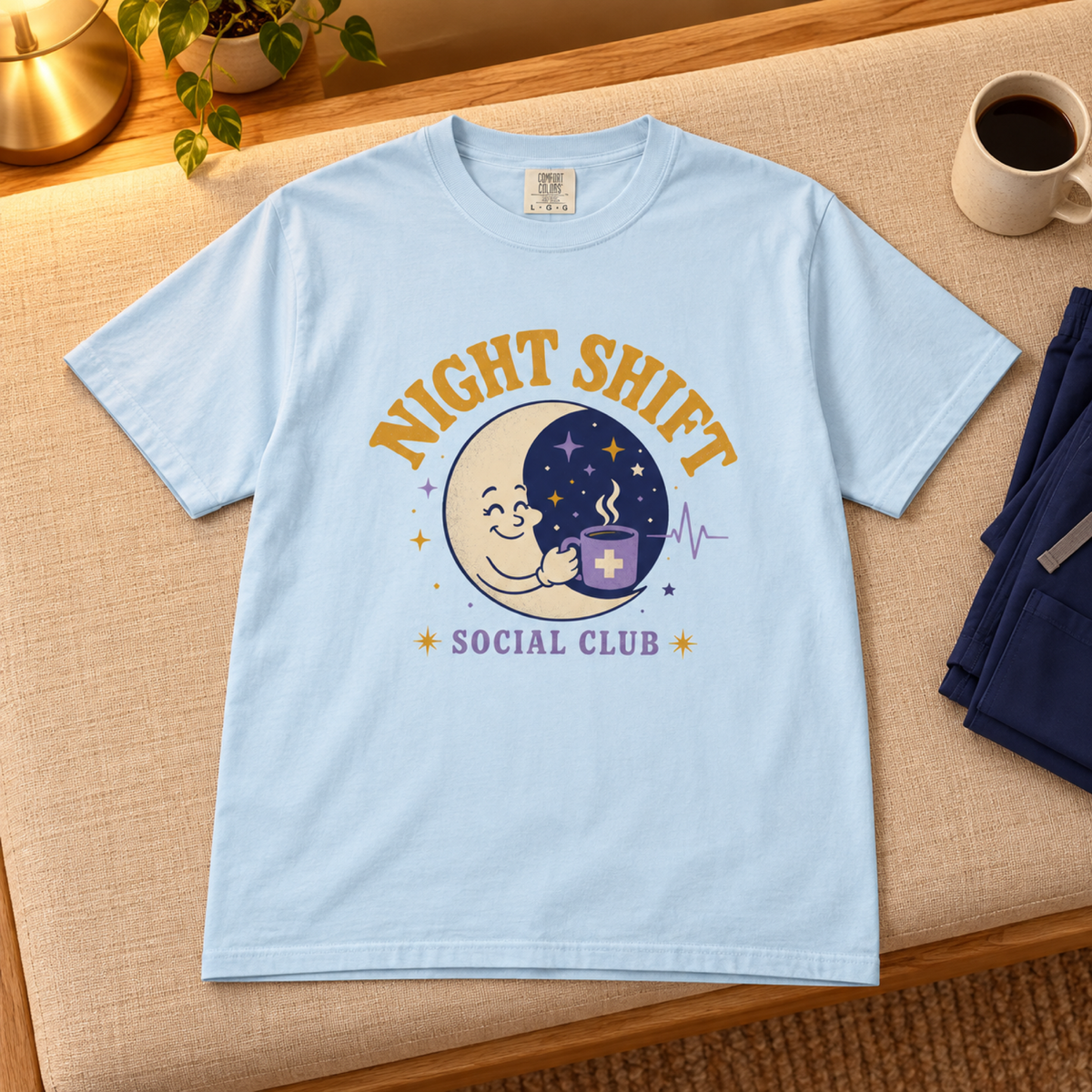 Chambray blue Night Shift Social Club tee flat lay on a warm linen bench in a cozy evening staff lounge