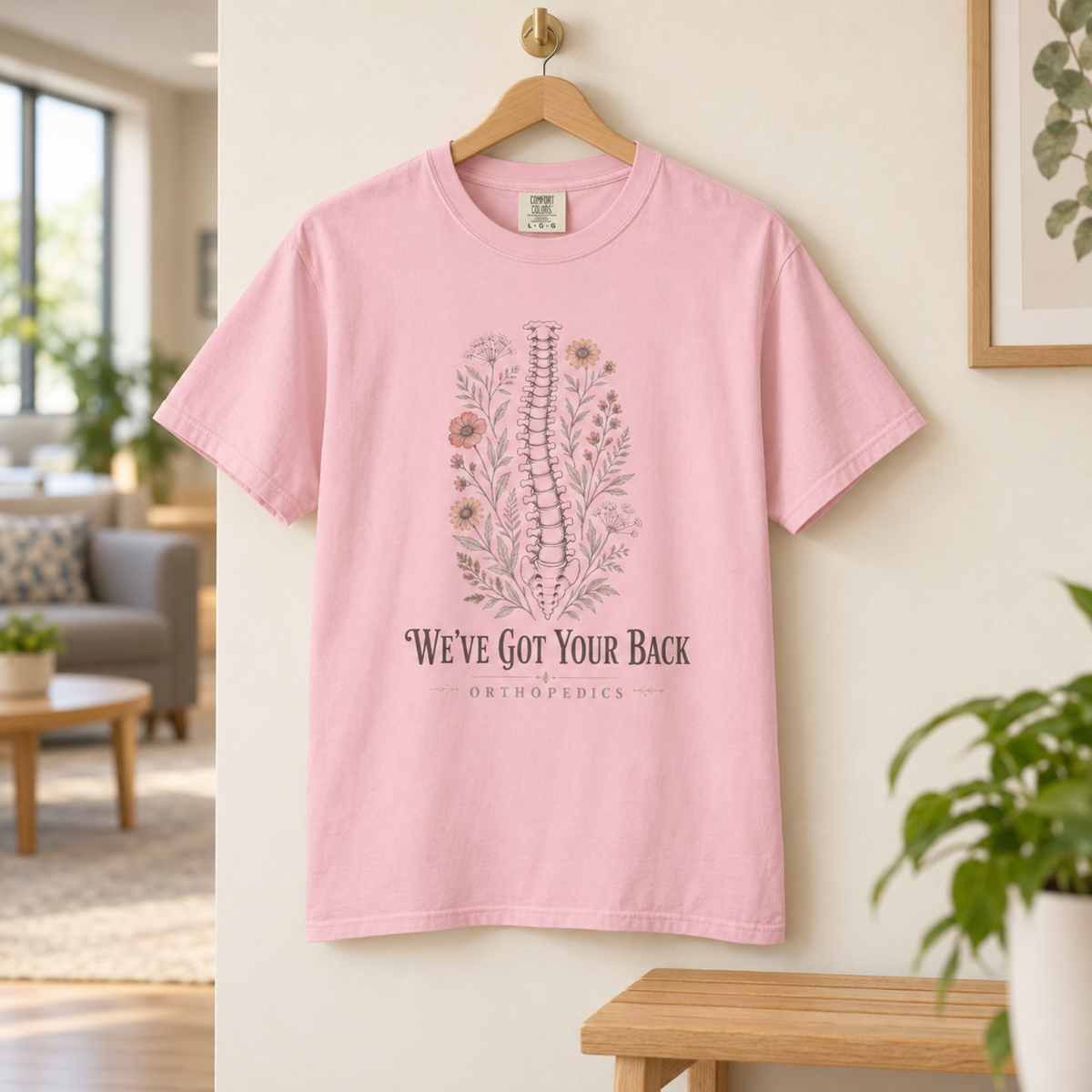 Blossom pink We've Got Your Back Orthopedics floral spine tee on an oak hanger in a warm clinic staff lounge