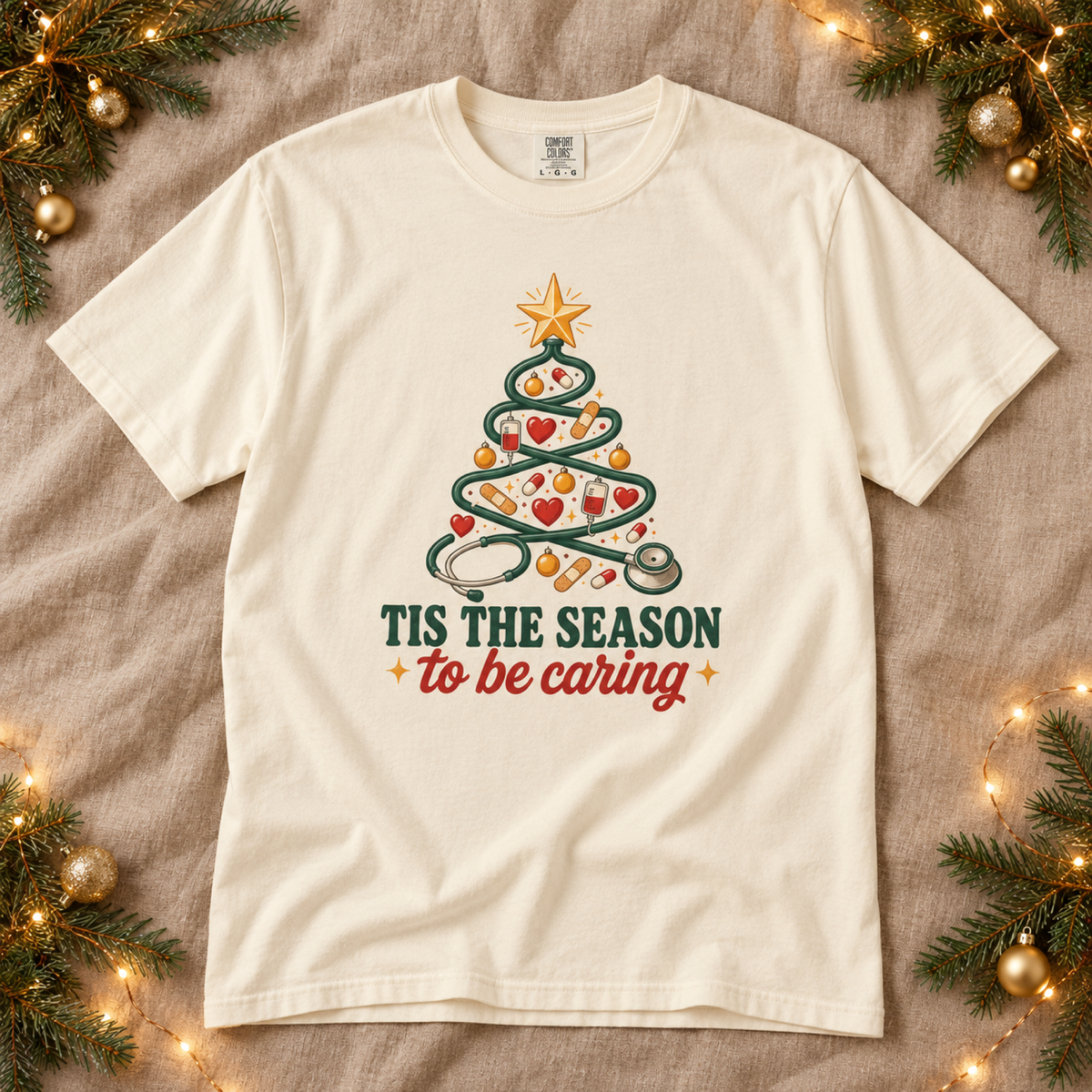Ivory Tis the Season to Be Caring Christmas tee flat lay on taupe linen with pine and gold ornaments